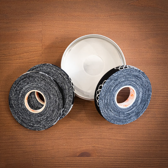 3 rolls of ClimbSkin Tape of varied thickness in black with white spiral design. Laid on top of one another on wooden surface with metal tin.