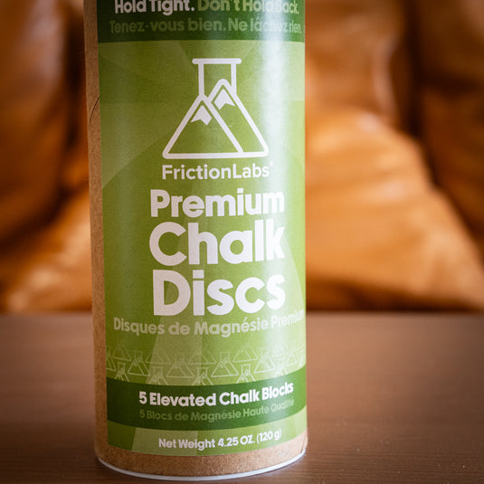 A cylindrical container of FrictionLabs Premium Chalk Discs, labeled in green, on a wooden surface. Contains five chalk blocks. A close up image.