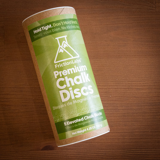 A cylindrical container of FrictionLabs Premium Chalk Discs, labeled in green, on a wooden surface. Contains five chalk blocks.