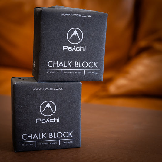 Two blocks of Psychi Chalk in black square paper packaging, dispalyed on a wooden surface. One block is balancing on the top of another block.