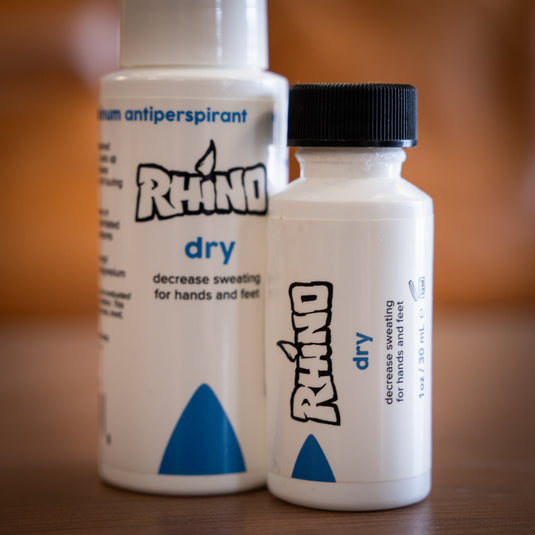 Two bottles of Rhino Skin Dry - decrease sweating for hands and feet. One large spray bottle and one smaller brush on applicator bottle. Both in white and dark blue design. Displayed on wooden surface.