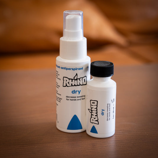 Two bottles of Rhino Skin Dry - decrease sweating for hands and feet. One large spray bottle and one smaller brush on applicator bottle. Both in white and dark blue design. Displayed on wooden surface.
