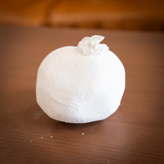 Ball of Chalk in white netting material. Displayed on wooden surface with a few specs of chalk on the surface.