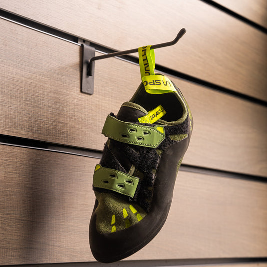 A climbing shoe, La Sportiva Tarantula velcro in olive neon hanging on a black hook against a textured metallic background.