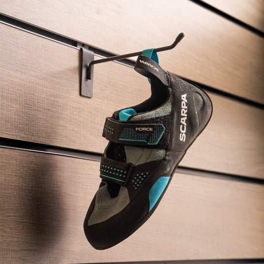 A climbing shoe, Scarpa Force, hanging on a black hook against a textured metallic background.