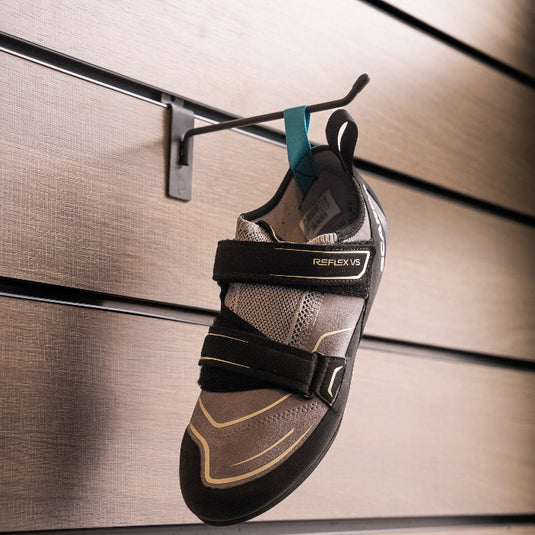 A climbing shoe, Scarpa Reflex VS WMN, hanging on a black hook against a textured metallic background.