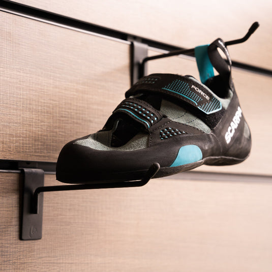 A climbing shoe, Scarpa Force, hanging on a black hook and with toe box balanced on another black hook against a textured metallic background.