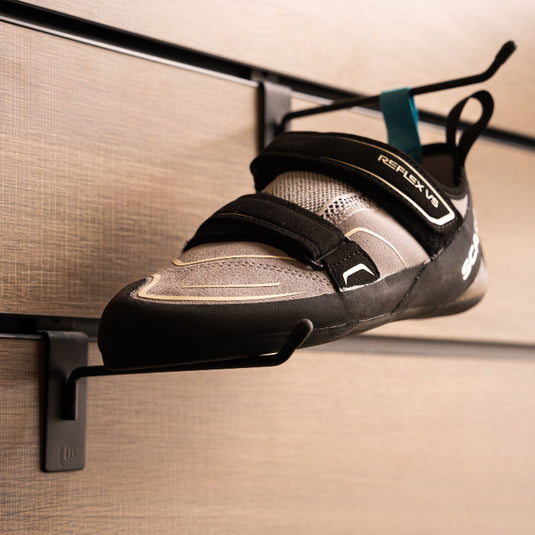 A climbing shoe, Scarpa Reflex VS WMN, hanging on a black hook and with toe box balanced on another black hook against a textured metallic background.