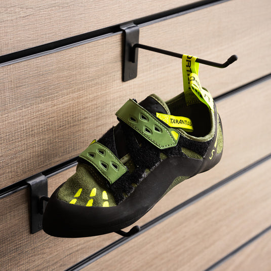A climbing shoe, La sportiva Tarantula on a black hook and with toe box balanced on another black hook against a textured metallic background.