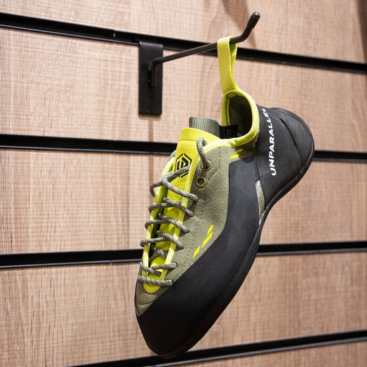 A climbing shoe, Unparallel Beat hanging on a black hook against a textured wooden background.