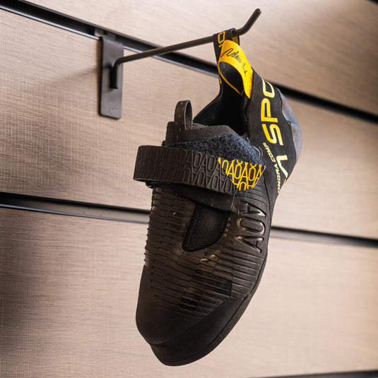 A climbing shoe, La Sportiva Ondra Comp, hanging on a black hook against a textured metallic background.