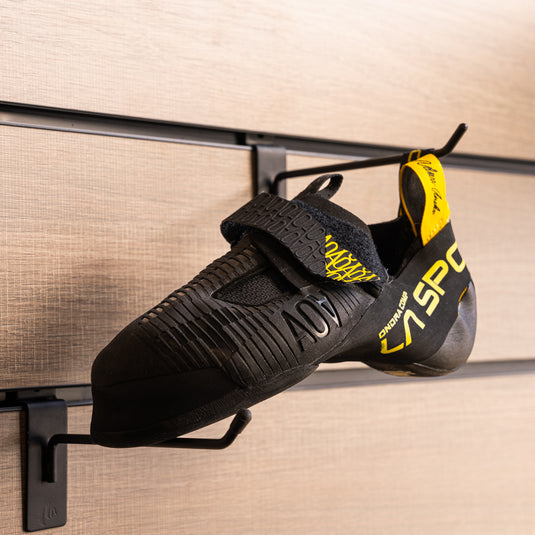 A climbing shoe, La sportiva Ondra Comp hanging on a black hook and with toe box balanced on another black hook against a textured metallic background.
