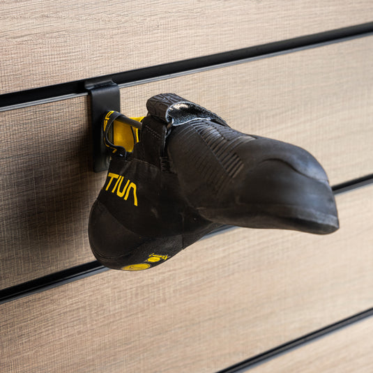 A La Sportiva ondra comp climbing shoe with a yellow and black heel strap loop, hanging on a wall-mounted holder against a textured background.