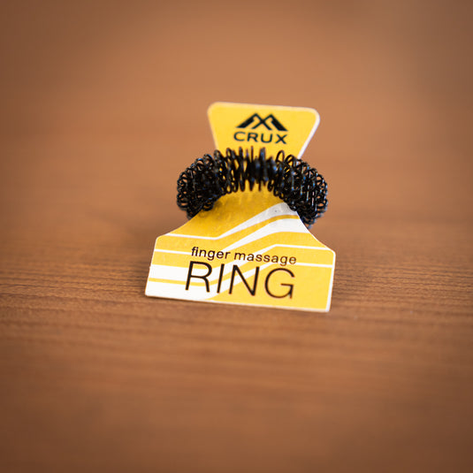 Close up of the Crux Finger Massage Ring in black. Displayed in yellow packaging and laid on wooden surface.