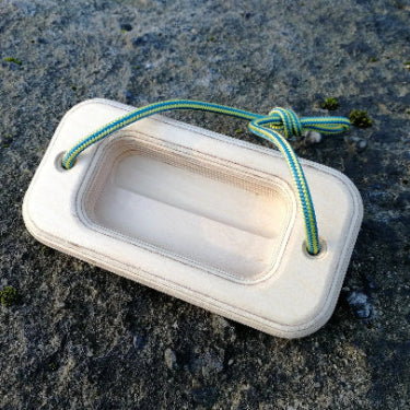 A wooden rectangular offcut handboard with a cut out in the centre. Green and yellow cord threaded through either side, resting on stone surface