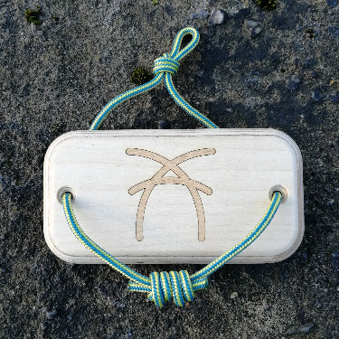 The back of a wooden rectangular offcut hangboard with green and yellow cord threaded through either side, with ash logo on the centre resting on stone surface