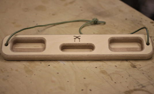 A wooden ash hangboard with various cutouts in the centre with blue and yellow cord threaded through either side. Displayed on wooden surface.