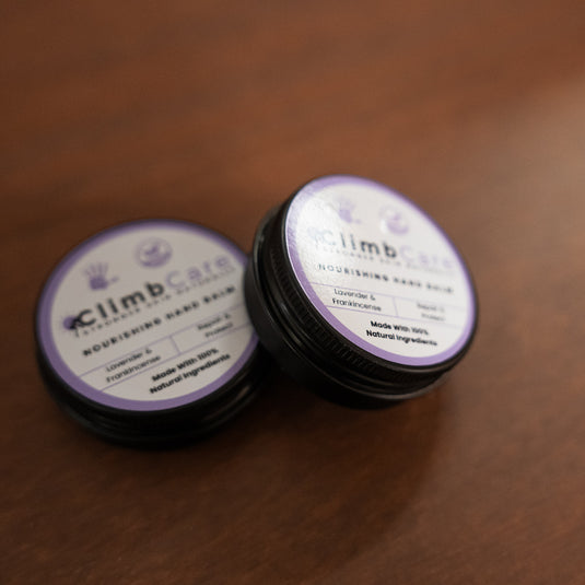 Two containers of ClimbCare nourishing hand balm laid on wooden surface in purple and white design. 